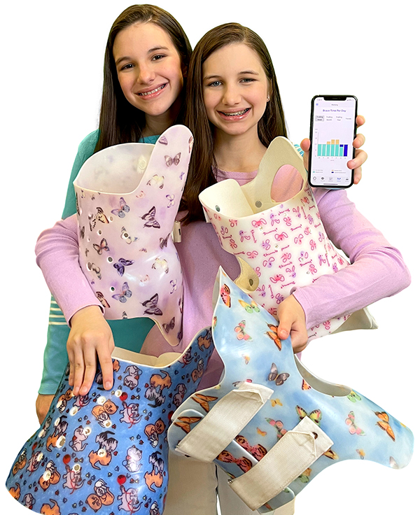 Hadley and Delany hold several beautifully decorated back braces while holding a phone with the BraceTrack app on the home screen.
